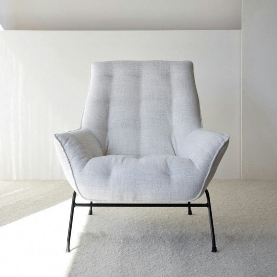 El Sillón de Diseño Tapizado en Mezclilla “Beige” es una silla moderna de respaldo alto y patas de metal negro, perfectamente integrada en el arte del diseño de interiores, descansando elegantemente sobre una alfombra suave y de color claro.