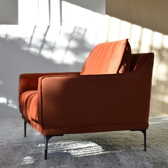 Un Sillón de Diseño en Terciopelo "Cuadrada" de color óxido con delgadas patas negras se encuentra sobre una alfombra gris claro en una habitación iluminada por el sol, proyectando sombras en la pared y el piso.