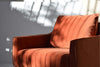 El Sillón de Diseño en Terciopelo "Cuadrada" en un vibrante color naranja se deleita con la luz del sol, su tono contrasta maravillosamente con sus patrones de sombras. Con elegantes patas de metal negro mate, combina elegancia y modernidad, invitándote a relajarte y descansar.