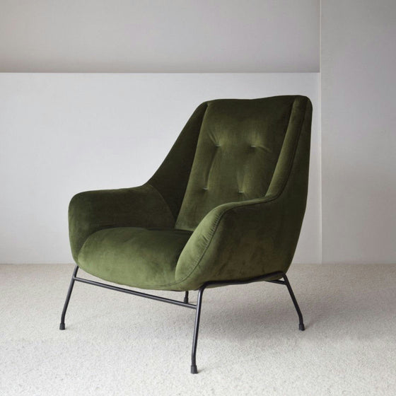El Sillón de Diseño en Terciopelo Verde "Oscuro" cuenta con un respaldo capitoné y se apoya elegantemente sobre patas de metal negro, elevando su decoración e interiorismo sobre un piso alfombrado claro.