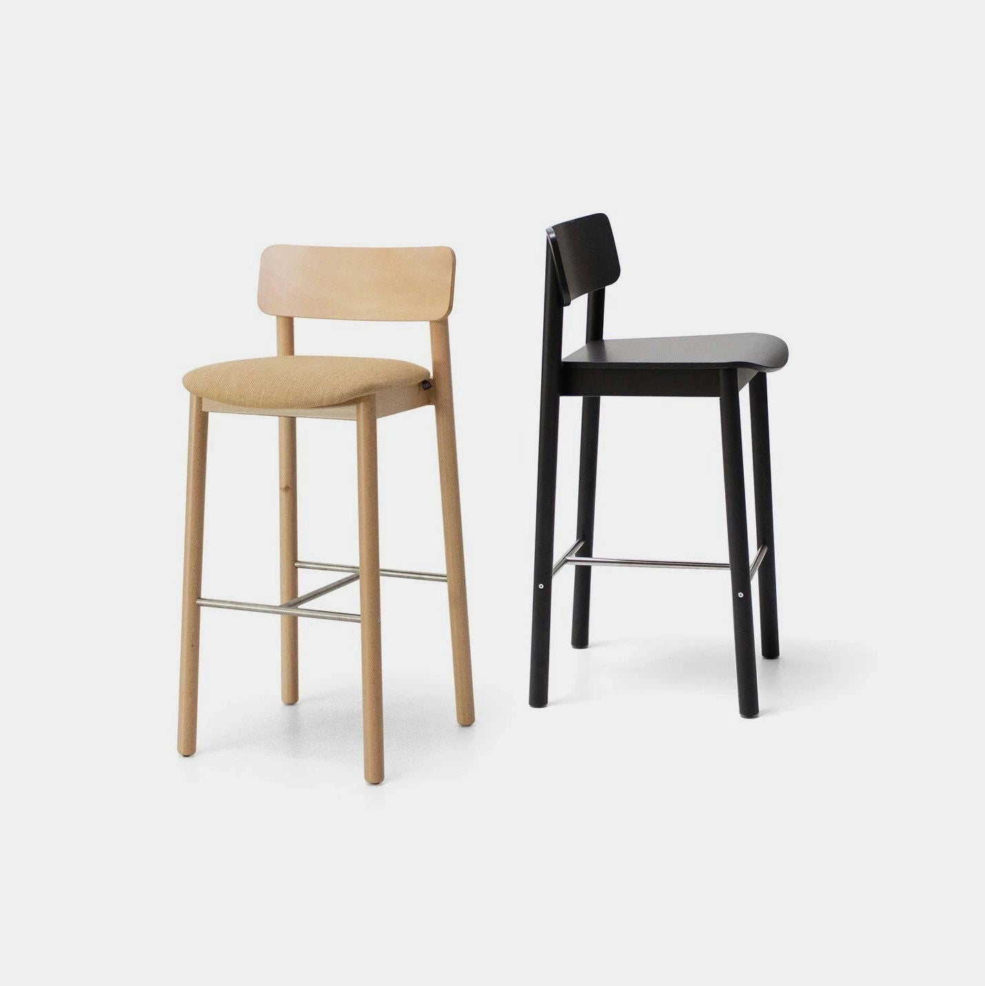 Dos Taburetes Altos en Madera y Tapizado Bion AT, uno con acabado en madera clara con un cojín color canela, el otro con acabado en madera oscura con un cojín negro, ofrecen una comodidad excepcional, gracias a sus reposapiés de metal, sobre un fondo liso.