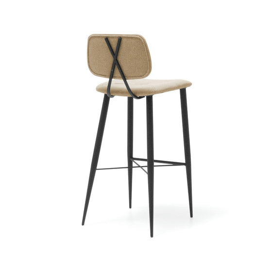 El Taburete Alto en Metal y Tapizado Umai es de color beige, presenta un respaldo rectangular con un diseño sofisticado y tiene patas de metal negro con un soporte en forma de X en el respaldo.