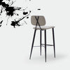 El Taburete Alto en Metal y Tapizado Umai presenta un asiento y respaldo en color beige con un diseño sofisticado, sostenido por elegantes patas de metal negro. Está ubicado sobre un fondo blanco con artísticas salpicaduras de tinta negra que adornan la esquina superior izquierda.