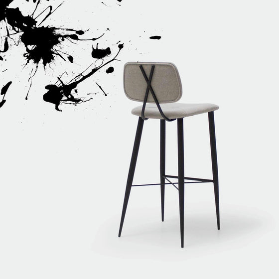 El Taburete Alto en Metal y Tapizado Umai presenta un asiento y respaldo en color beige con un diseño sofisticado, sostenido por elegantes patas de metal negro. Está ubicado sobre un fondo blanco con artísticas salpicaduras de tinta negra que adornan la esquina superior izquierda.