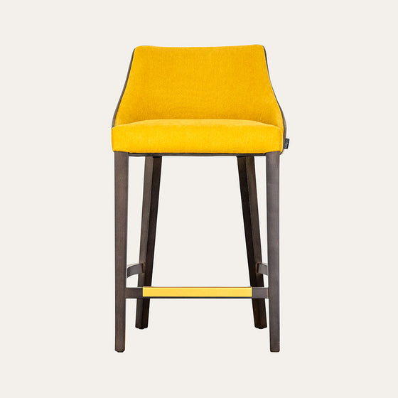 Un Taburete en Madera y Tapizado Xally, que presenta un asiento y respaldo acolchados de color amarillo personalizables, sostenido por patas de madera oscura y visto desde el frente. Este taburete en madera se integra a la perfección en cualquier diseño contemporáneo.