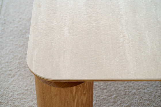 La Mesa de centro cuadrada en cerámico DM y fresno natural Línea, perfecta para un salón contemporáneo, presenta un diseño rectangular de color claro con bordes redondeados y patas cilíndricas gruesas, agregando elegancia a cualquier decoración del hogar sobre un piso alfombrado.