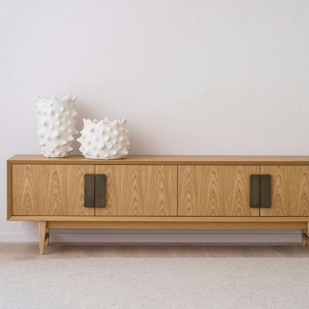 El Mueble TV en DM y roble roble y latón envejecido Oia tiene tiradores verdes y complementa una pared blanca inmaculada. Encima se sitúan dos jarrones de cerámica blanca texturizada y debajo una alfombra beige que realza su diseño minimalista.