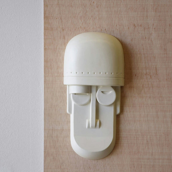 Una Figura Máscara Decorativa en Cerámica Blanco mate está montada en una pared de madera con una elegante franja vertical blanca, capturando la esencia del diseño contemporáneo.