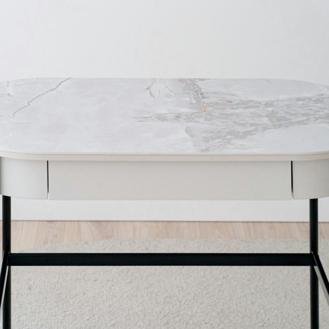 El Escritorio Ovalado en Mármol Cerámico Blanco N.26 (Gris arena) destaca por su diseño contemporáneo con cubierta de mármol cerámico claro, cajón blanco y patas negras, colocado sobre alfombra clara junto a una pared blanca.