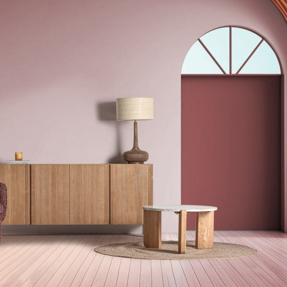 Una moderna sala de estar cuenta con un elegante aparador de madera DM (180x45x80 cm) Copenhague. Una lámpara sobre la mesa de centro redonda reposa sobre una alfombra, con una ventana en forma de arco que realza las paredes rosas y el suelo de madera clara, creando un toque de diseño moderno.