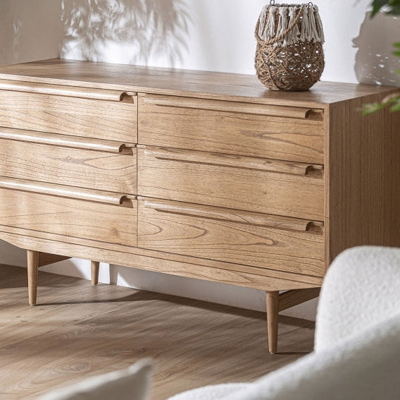 El Aparador de madera mindi (150 x 45 cm) Nyry, con seis cajones, se coloca contra una pared blanca, representando el mobiliario contemporáneo. Sobre él se encuentra una cesta tejida decorativa con tapa para un almacenamiento eficiente, mientras que las sombras de las hojas realzan la estética serena.