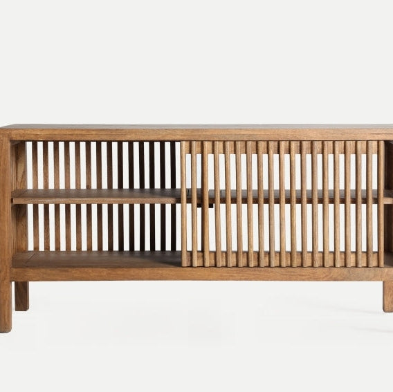 El Aparador de madera en mango (121x36x61 cm) Trevoux es una mesa consola que presenta un elegante diseño de listones verticales, estantes abiertos en un lado y una puerta corrediza de listones, todo elaborado con madera de mango de primera calidad para incorporar el estilo nórdico sobre un fondo liso.
