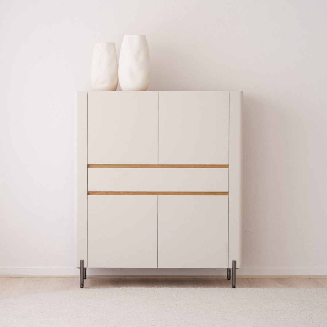 Un ARMARIO minimalista. ARENA MATE Y ROBLE NATURAL con dos jarrones blancos en la parte superior se destaca contra una pared blanca, resaltando su DISEÑO ATEMPORAL, mientras que cortinas transparentes enmarcan elegantemente el lado izquierdo.