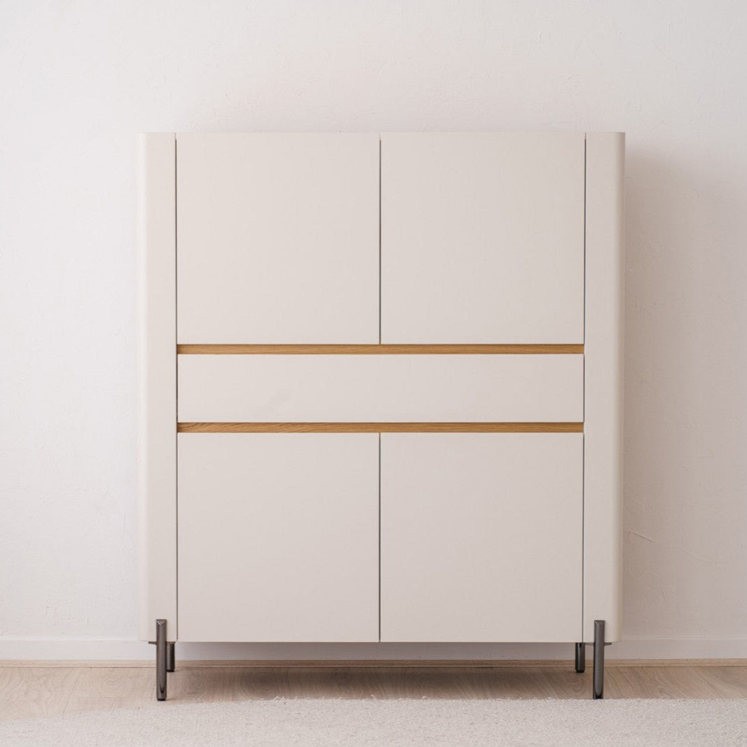 El ARMARIO. ARENA MATE Y ROBLE NATURAL es una pieza minimalista con cuatro puertas y dos líneas de acento de roble natural, colocadas contra una pared blanca lisa, ejemplificando un diseño atemporal.