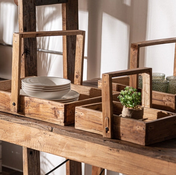 Sobre una mesa de madera rústica se encuentran Cajas en madera de caoba Akviran (3 piezas); una sostiene platos blancos apilados, mientras que otra muestra una pequeña planta en maceta y tazas de vidrio.
