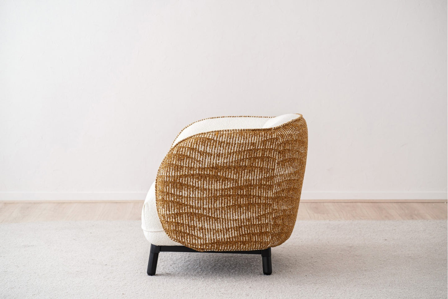 El Sillón curvo tapizado en blanco bouclé y mostaza CURVA (85×84×77 cm) se muestra de espaldas sobre una alfombra clara en una habitación minimalista con suelo de madera y paredes blancas.
