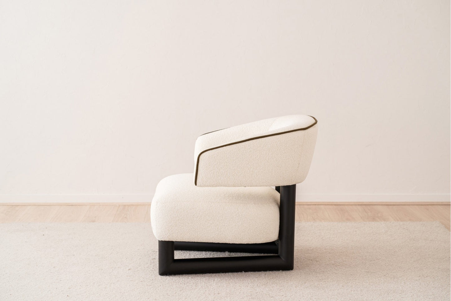 Butaca de diseño tapizada en tela bouclé blanco roto y madera negra NEO, vista lateral sobre alfombra clara en ambiente minimalista con paredes pálidas y zócalo de madera.