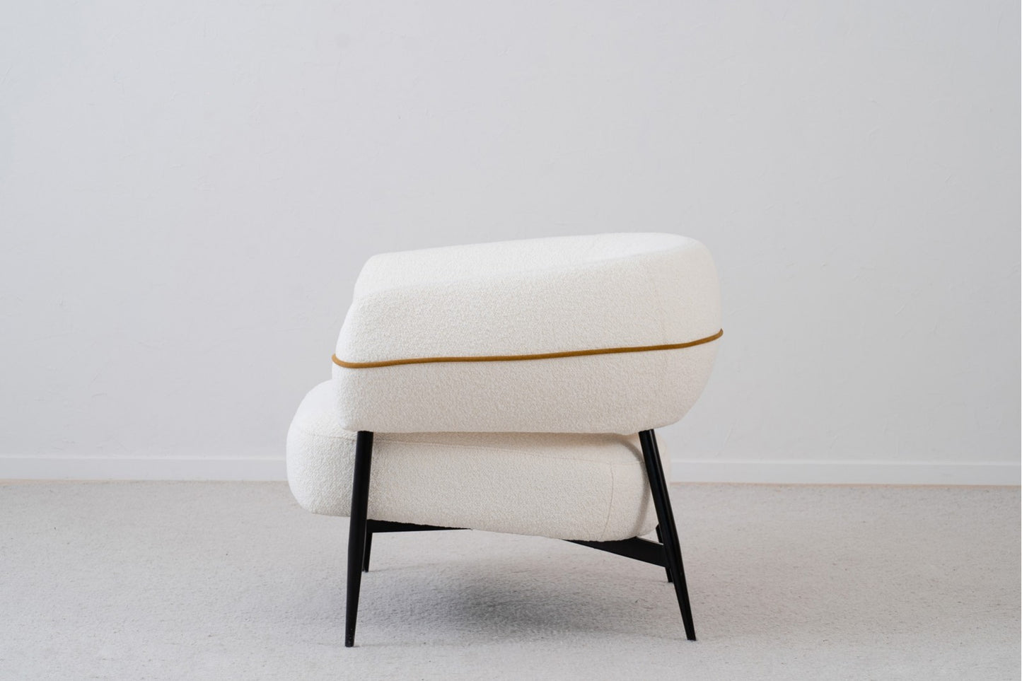 Un sillón curvo KYRTOS contemporáneo con tapicería de bouclé blanco roto, ribete ocre y patas de metal negro (89×84×74 cm), expuesto de lado contra una pared blanca lisa y suelo enmoquetado.