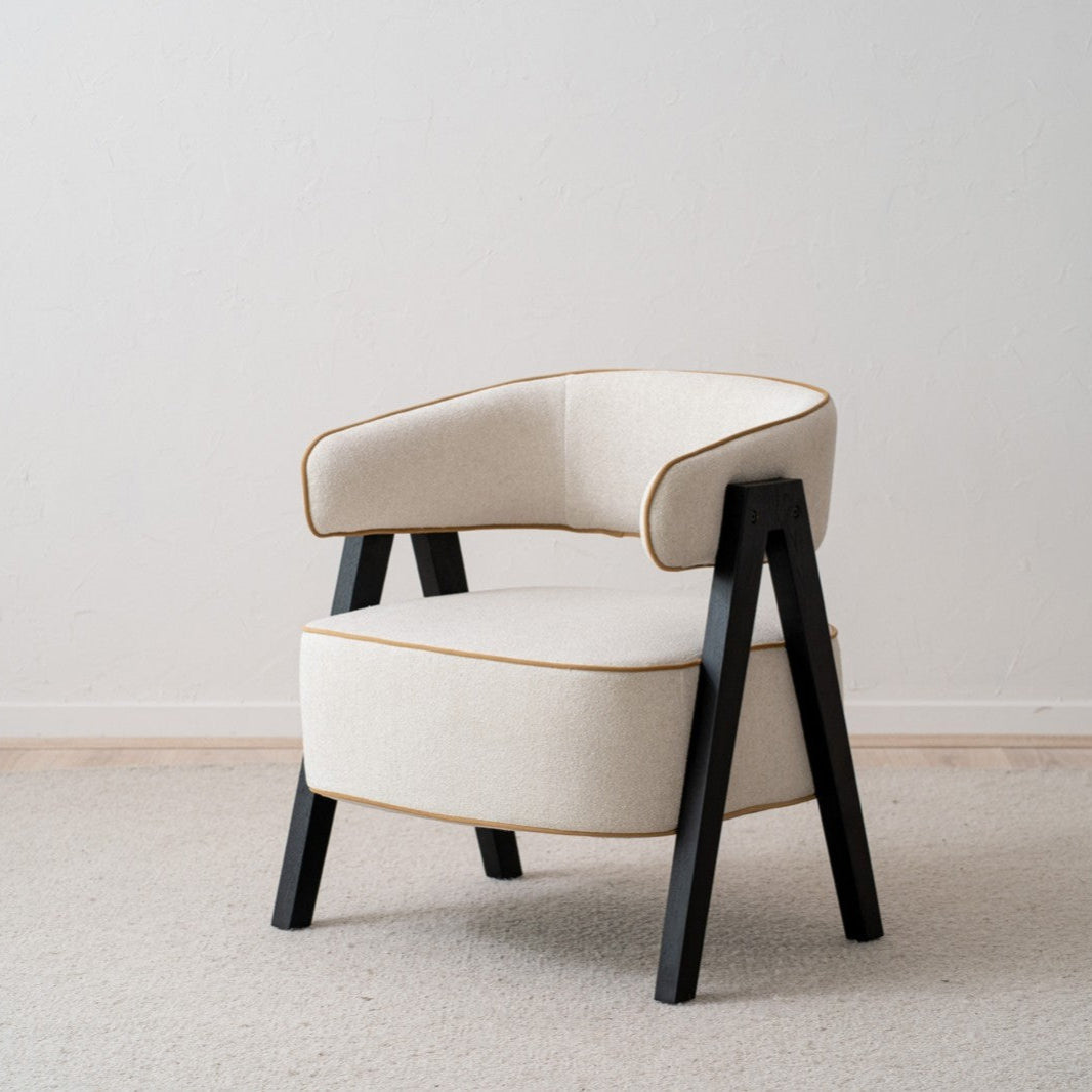 El sillón tapizado en beige y madera de fresno negro Capri, con un asiento acolchado en color crema y elegantes patas de madera negra, está colocado sobre una alfombra clara contra una pared blanca sencilla, agregando un aire de estilo moderno a la habitación.