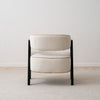 El sillón tapizado en beige y madera de fresno negro Capri, con su diseño moderno y asientos acolchados, reposa elegantemente sobre una alfombra de tonos claros contra una pared neutra.