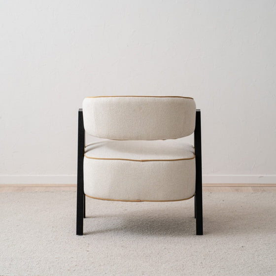 El sillón tapizado en beige y madera de fresno negro Capri, con su diseño moderno y asientos acolchados, reposa elegantemente sobre una alfombra de tonos claros contra una pared neutra.