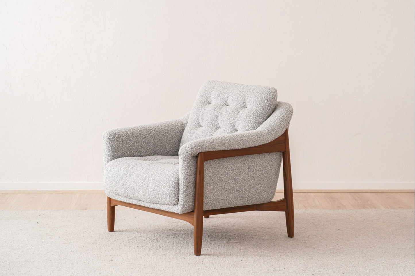 A conjunto de butaca y puff de madera maciza nogal y tela bouclé blanco roto - mid century - FYFTY se exhibe sobre una alfombra beige en una sala minimalista con suelo de madera clara y pared blanca texturizada.