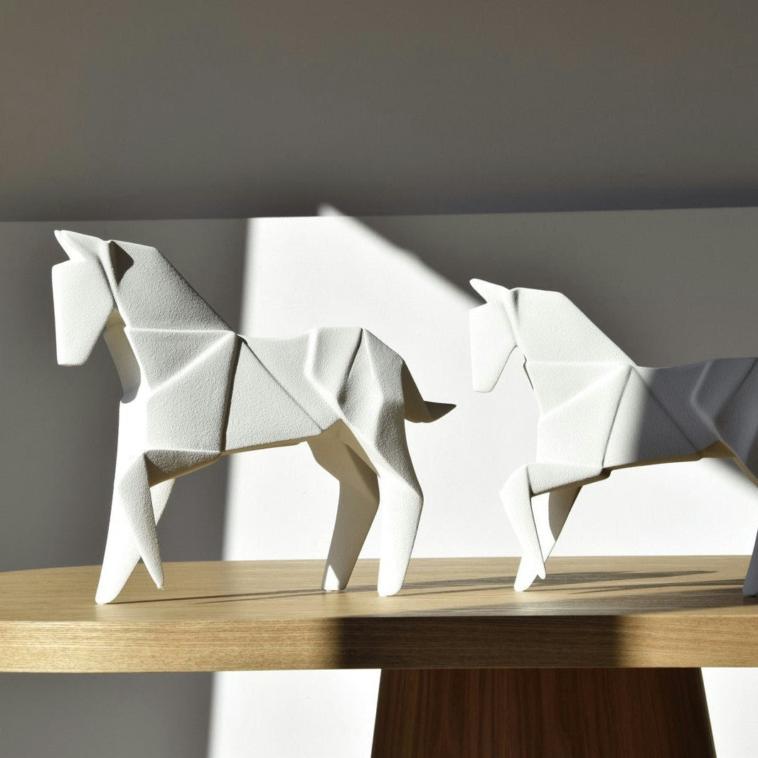 Dos caballos de cerámica blanca, parecidos a las "Figuras decorativas en cerámica Blanco mate Caballo Origami", se encuentran sobre un estante de madera iluminado por la luz del sol.
