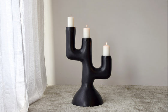 Un Candelabro Totem N.1 Metal Negro 3 Velas, un candelabro negro con forma de cactus, con tres velas blancas encendidas, se encuentra sobre un piso alfombrado cerca de una cortina blanca, mostrando una artesanía única y un diseño contemporáneo.