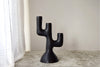 Una escultura abstracta de cactus negra con tres brazos, similar al Candelabro Totem N.1 Metal Negro 3 Velas, se encuentra sobre una alfombra gris frente a una pared pálida y una cortina blanca, encarnando el diseño contemporáneo.