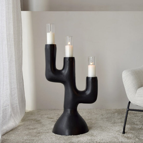 El Candelabro Totem N.1 Metal Negro 3 Velas, un candelabro escultural inspirado en un cactus, muestra tres velas encendidas y está elegantemente posicionado en el piso cerca de un sillón beige y una cortina blanca, realzando el espacio con un toque de diseño contemporáneo.