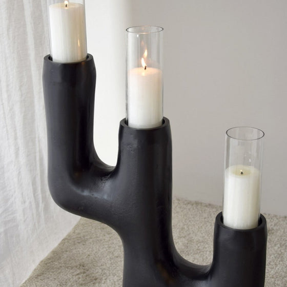 El Candelabro Totem N.1 Metal Negro 3 Velas es un candelabro escultórico negro que sostiene tres velas blancas en cilindros de vidrio sobre un piso alfombrado: una exquisita pieza de artesanía única que muestra el diseño contemporáneo.