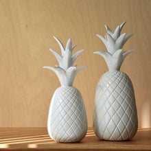  La colección Figuras Decorativas en Cerámica Blanca Piña incluye dos esculturas de piña de cerámica en varios tamaños sobre una superficie de madera, resaltando el exquisito diseño artesanal sobre un fondo de madera clara.