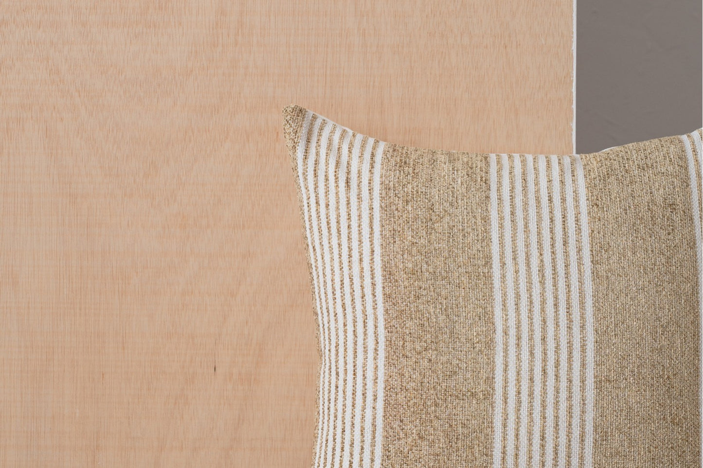 El Cojín de algodón con franjas blanco y camel 50X50CM - natural - ERO N3 se sitúa delante de un panel de madera clara, con una pared gris a la derecha.