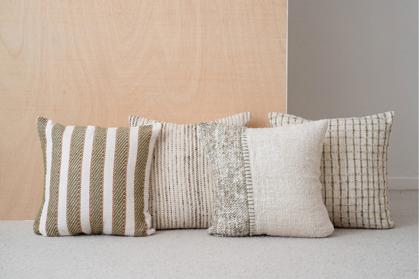 Cuatro cojines decorativos, entre ellos el cojín BERTE N4 de rejilla de algodón blanco, verde y beige (50x50 cm), se disponen en fila sobre un fondo de madera clara y pared blanca.