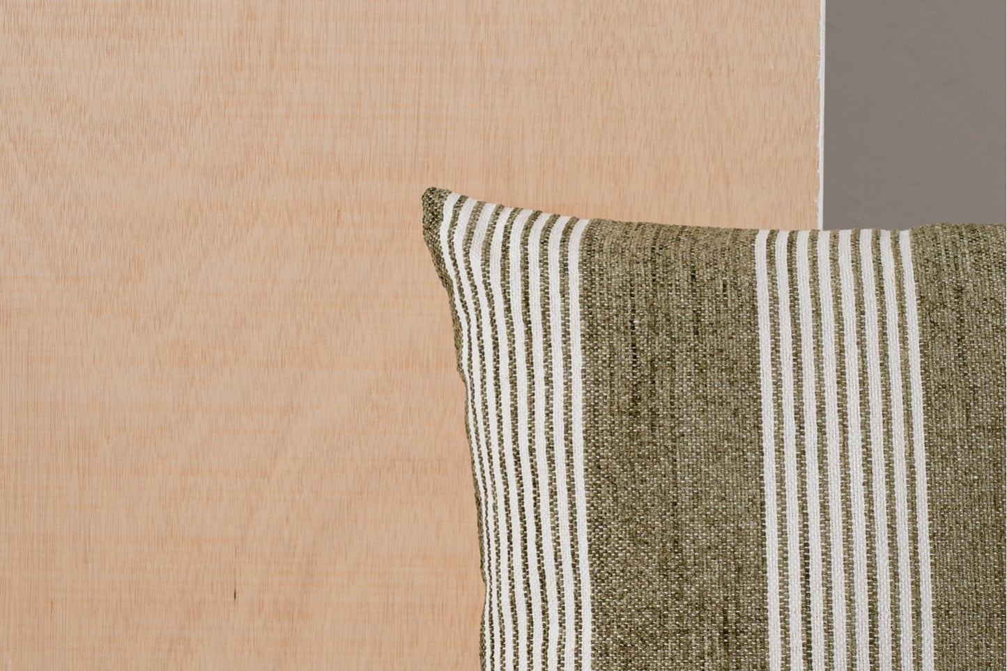 El Cojín de algodón blanco y verde oliva con franjas 50x50cm BERTE N5 se apoya sobre una superficie de madera clara, junto a una pared gris visible a la derecha.