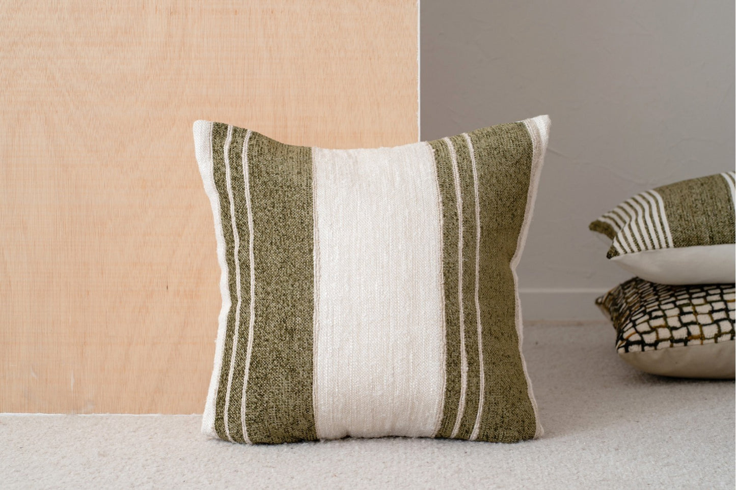 El cojín de algodón blanco, beige y verde oliva con franjas 50x50cm - natural - BERTE N6 descansa sobre una superficie de madera, con dos cojines decorativos apilados al fondo sobre un suelo enmoquetado.