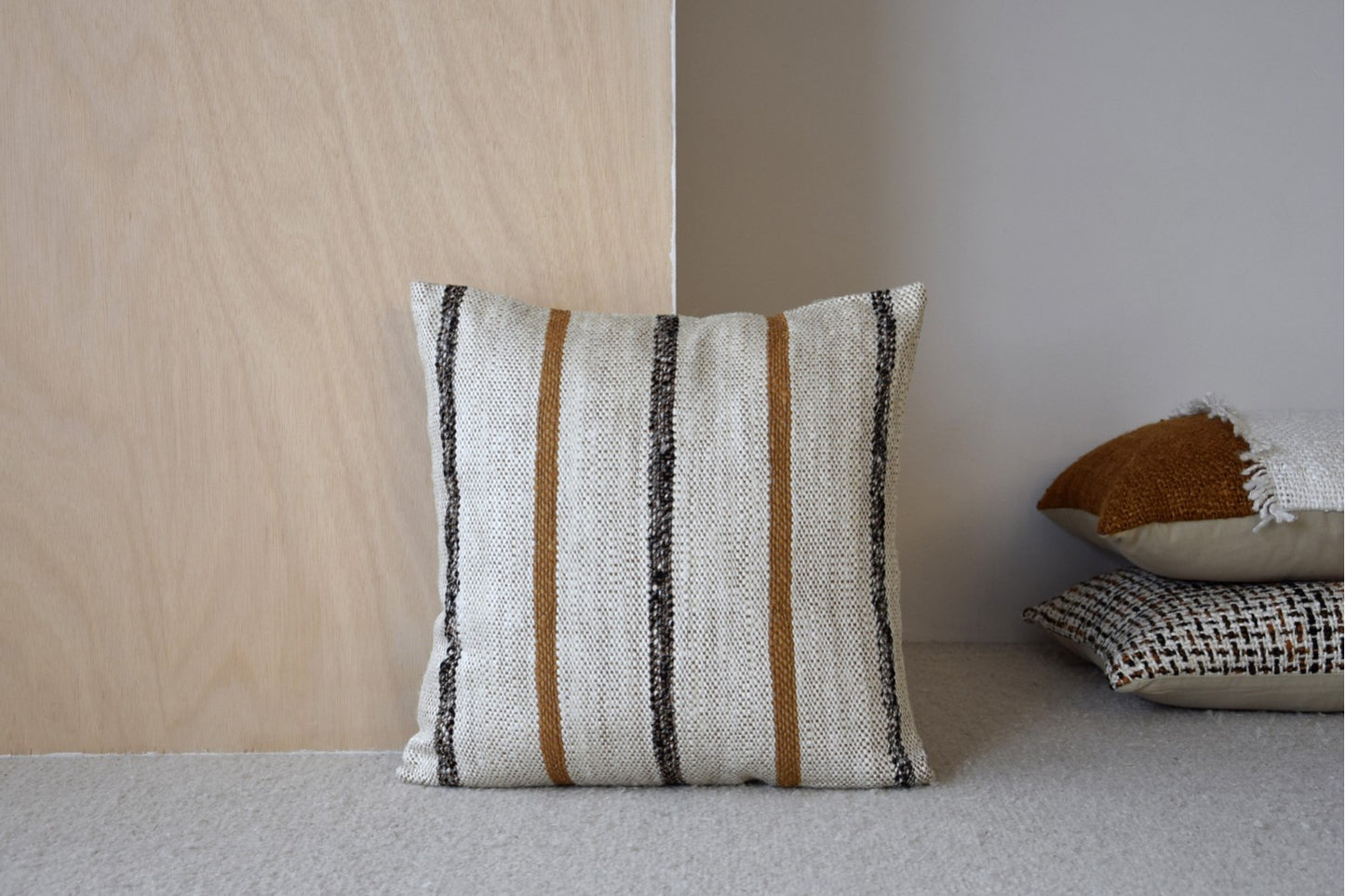 A Cojín de algodón mostaza y blanco con líneas estrechas 50x50cm - natural - LAZA N6 descansa sobre un panel de madera clara; otros dos cojines están apilados cerca, sobre el suelo enmoquetado.
