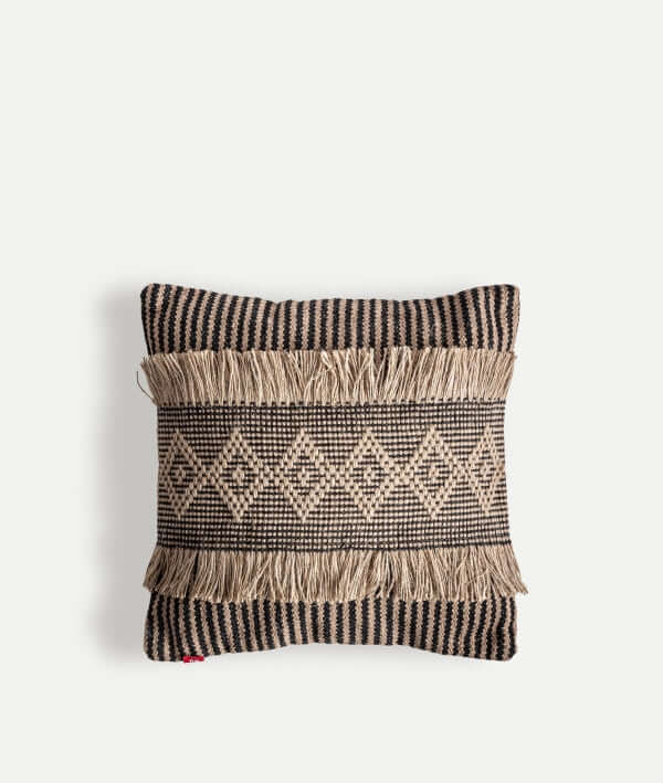 El cojín KODU BLAHIR de yute marrón claro y poliéster negro (45×45 cm) presenta un diseño boho con patrón geométrico, flecos horizontales y bordes a rayas, sobre fondo blanco.