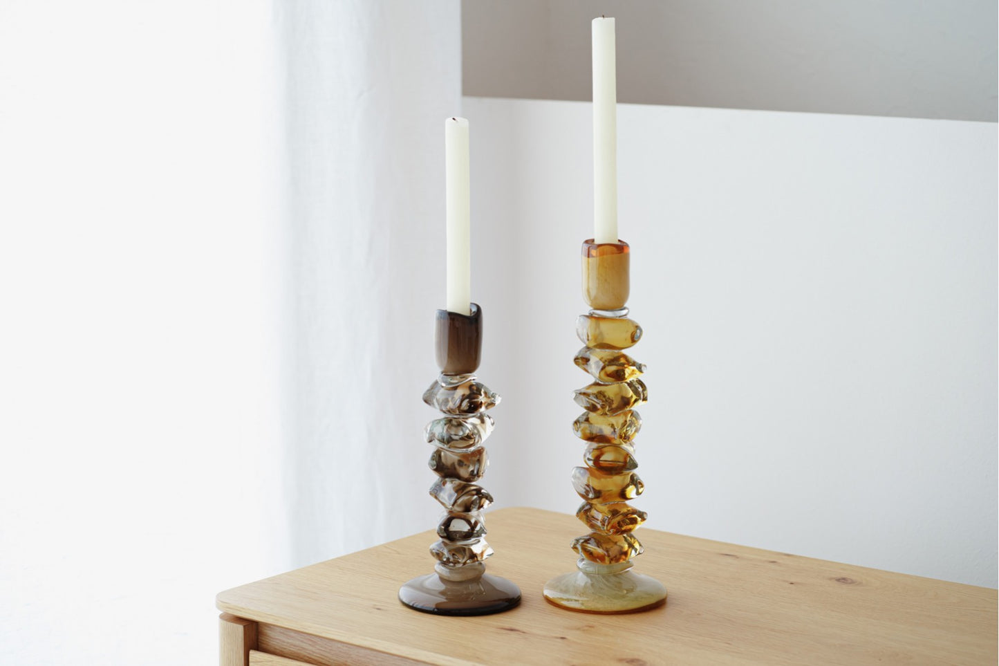 El Set de Candelabros de vidrio verde oliva / ámbar - ALDAN con velas blancas decora una superficie de madera frente a un fondo blanco y gris, ideal para la decoración del hogar.