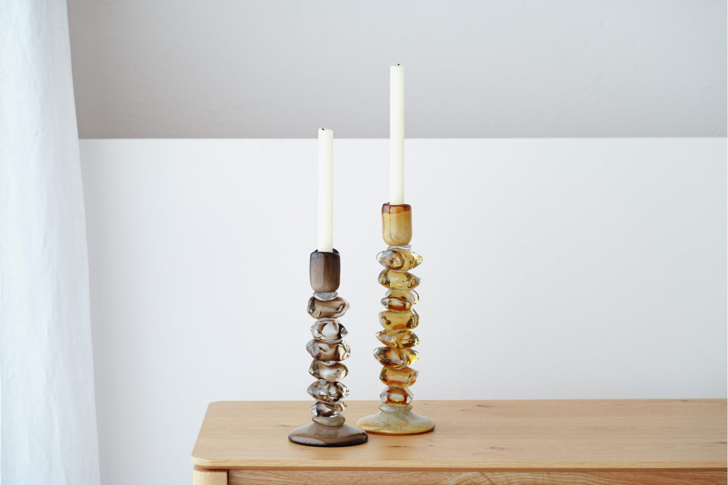 El set de candelabros de vidrio verde oliva / ámbar ALDAN se exhibe sobre una superficie de madera, aportando un toque elegante a la decoración del hogar frente a una pared blanca.