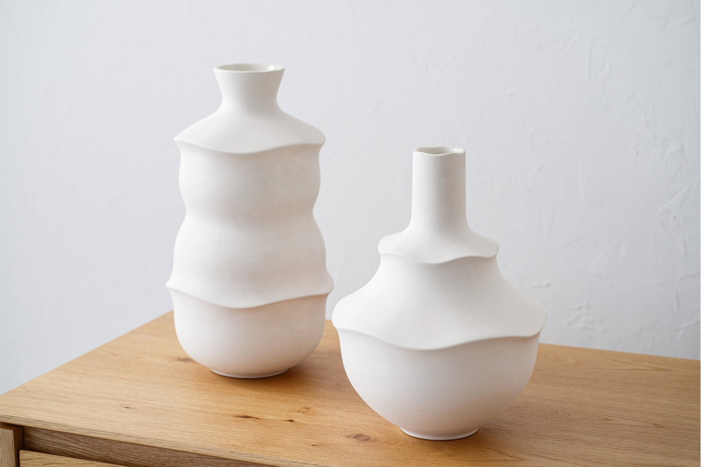 El set de jarrones de cerámica blanco mate alto y bajo ALEN, con diseño orgánico, aporta estilo único a la decoración interior sobre una superficie de madera clara junto a una pared blanca.