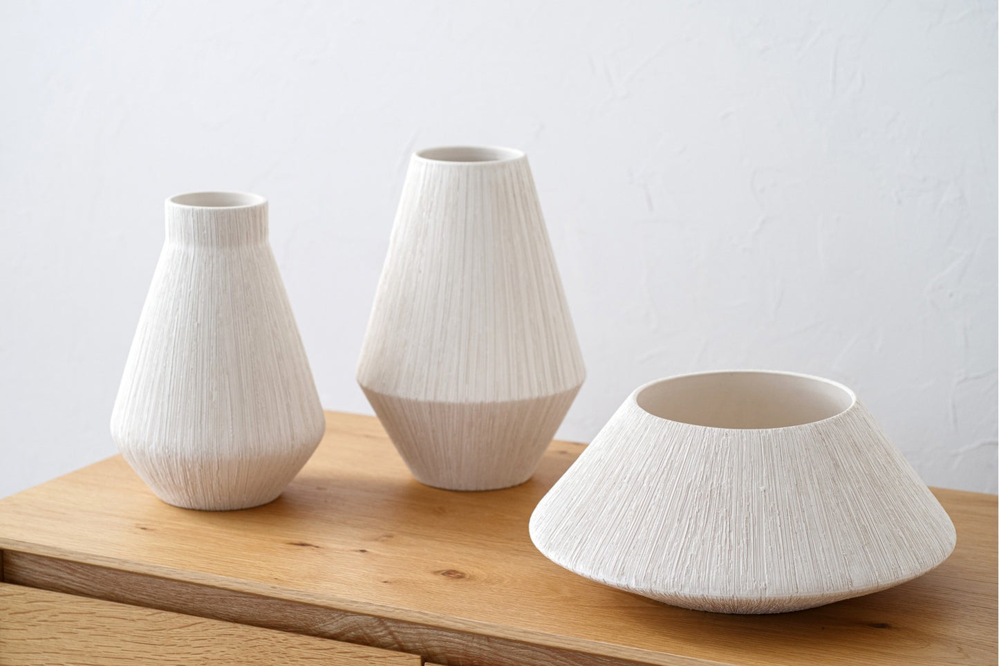El Set de jarrones y centro de mesa de cerámica blanca texturizada EINÉS, en color natural, decora una superficie de madera clara frente a una pared blanca, creando un elegante diseño artesanal.