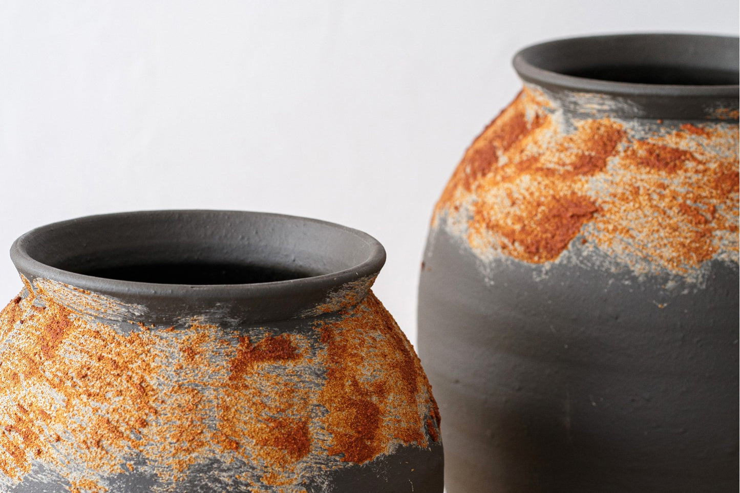 El Set de vasijas de cerámica negro óxido pequeña y grande FERRO son dos vasijas de cerámica rústica con textura de manchas de color óxido sobre arcilla gris, expuestas sobre un fondo blanco liso.