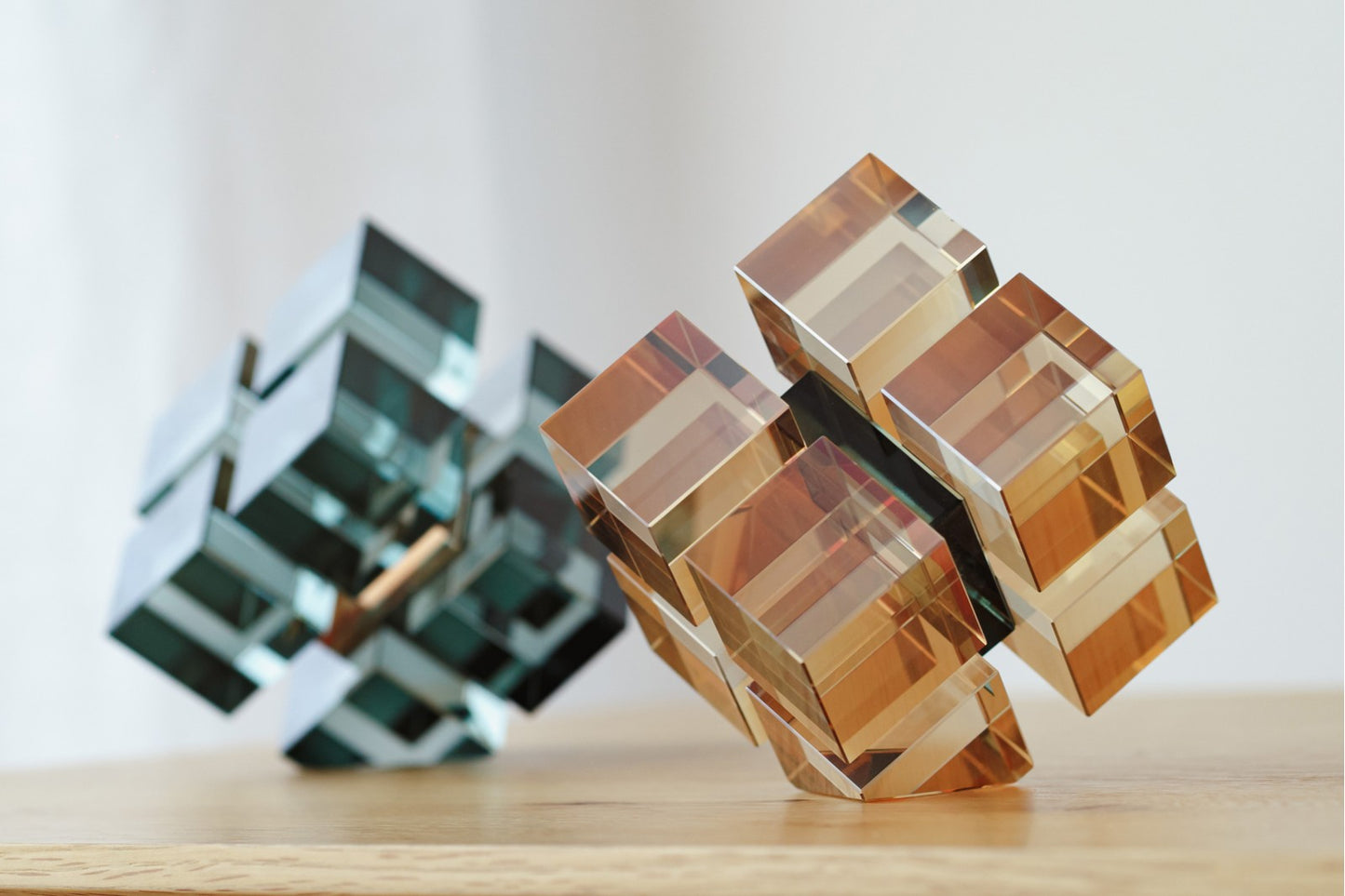 Dos piezas decorativas geométricas del set PUZZLE, en cristal turquesa y champagne, se exhiben sobre madera clara frente a un fondo neutro, aportando un toque moderno y elegante al ambiente.