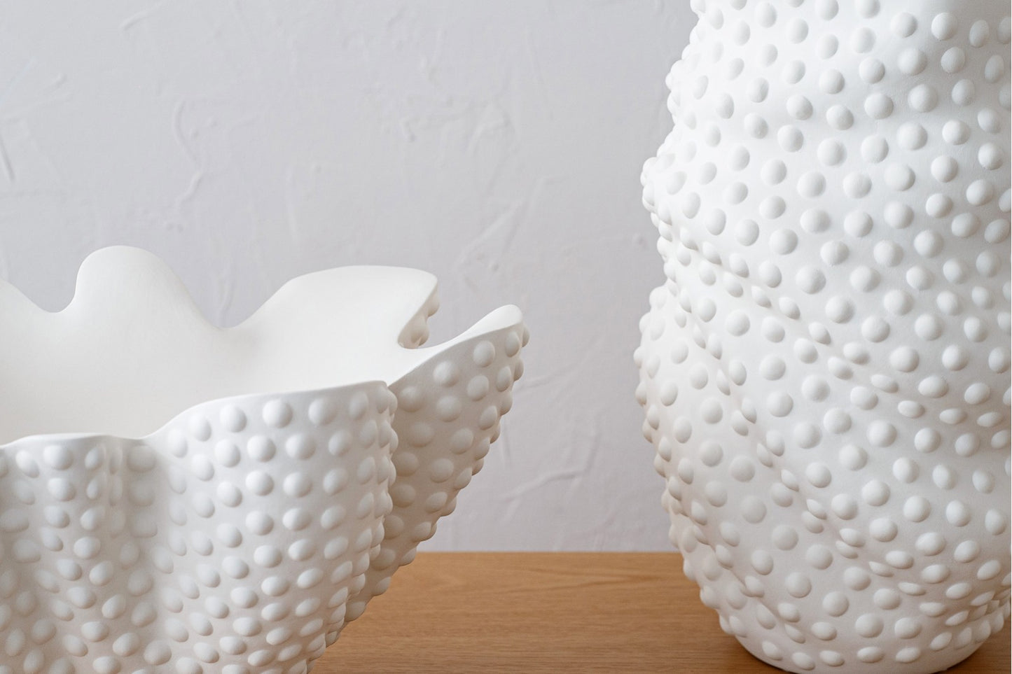 El set SOIA incluye un jarrón y un centro de mesa de cerámica blanco mate con textura, expuestos sobre una superficie de madera frente a una pared clara.