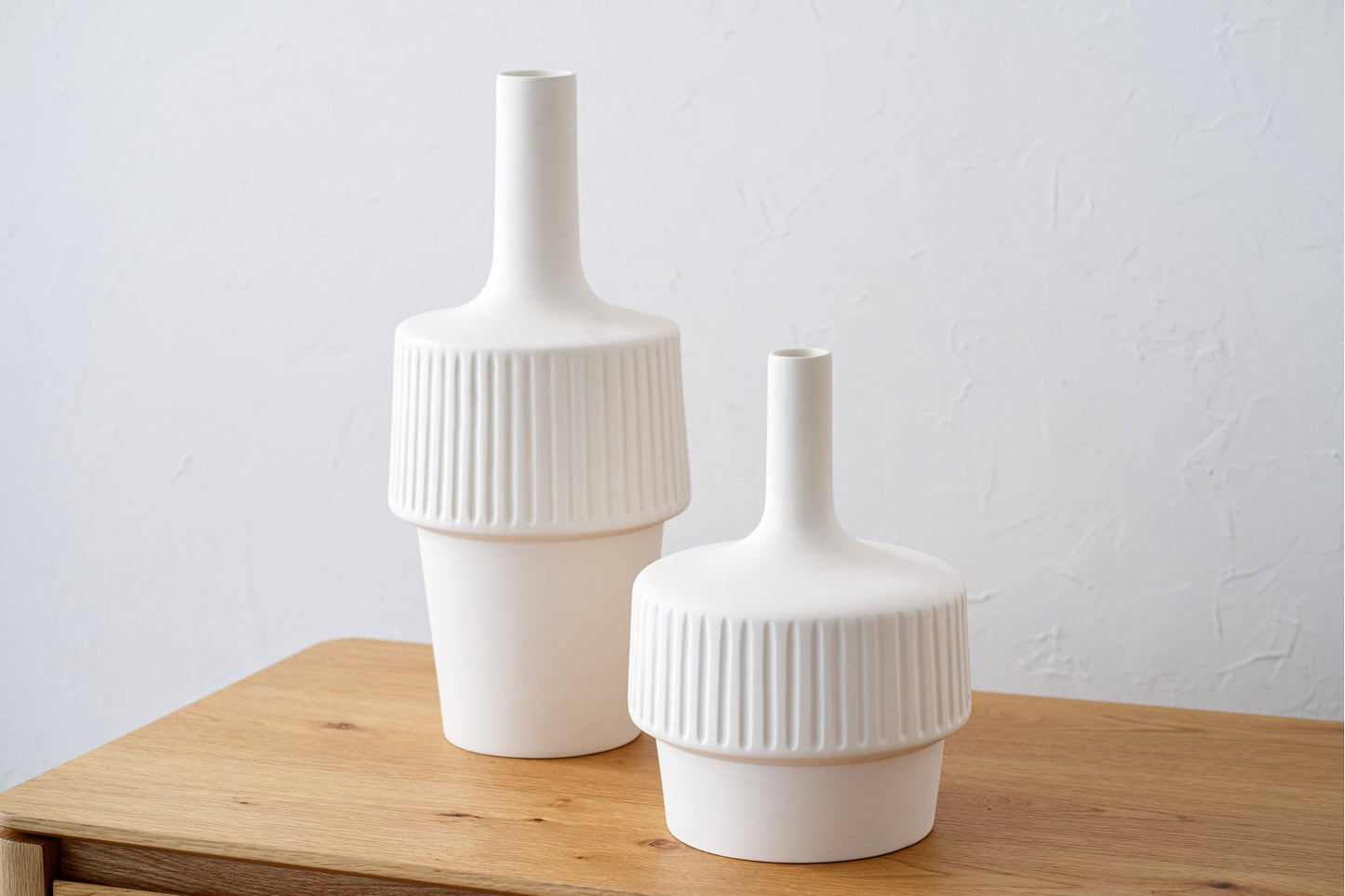 El set de jarrones de cerámica blanco mate grande y pequeño VESTA, con textura acanalada y cuello estrecho, destaca sobre madera clara y es ideal para decorar tu hogar frente a una pared blanca.