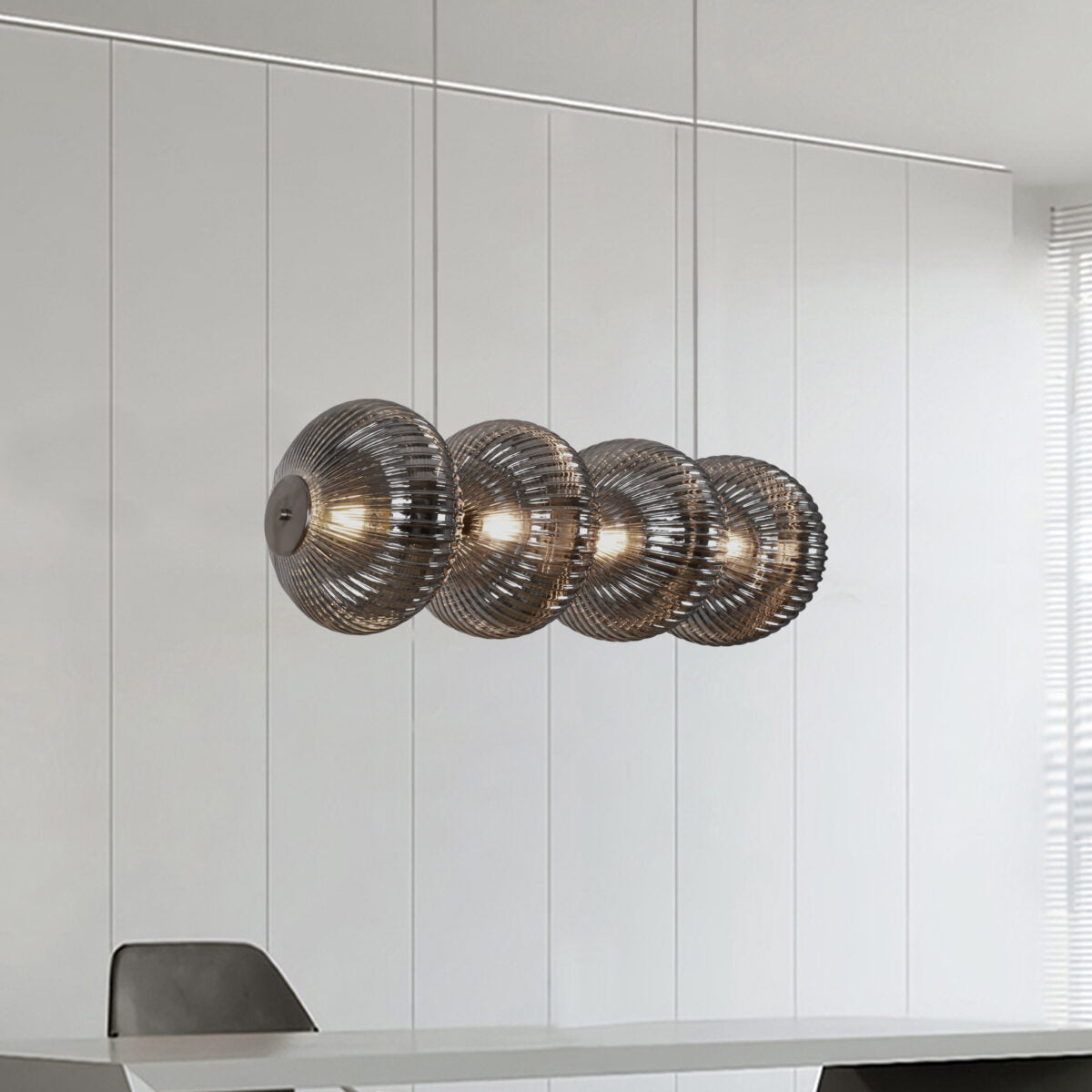 Un comedor moderno luce la elegante lámpara de techo S-Kairo 4, con cuatro pantallas esféricas de cristal. La decoración minimalista incluye una silla negra parcialmente visible, complementada con una cálida iluminación que crea un ambiente acogedor.