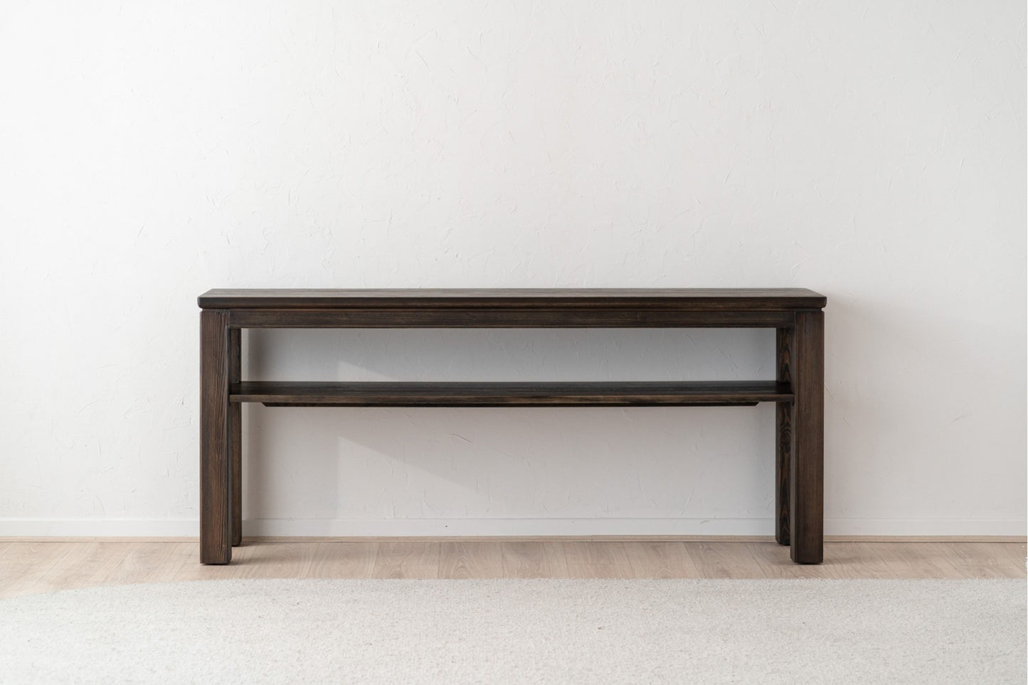 La Consola de madera maciza de pino teñido en oscuro 190×45×81 CM - contemporánea - IREA destaca por sus líneas simples y elegantes, ideal como mueble recibidor sobre un suelo claro junto a una pared blanca.