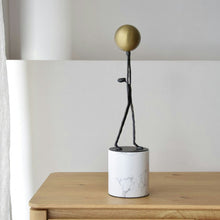  La Figura Decorativa en Mármol y Metal Equilibrio Celestial presenta una figura de palo minimalista que sostiene una esfera dorada sobre un cilindro de mármol blanco, perfecta como decoración lujosa en una mesa de madera.