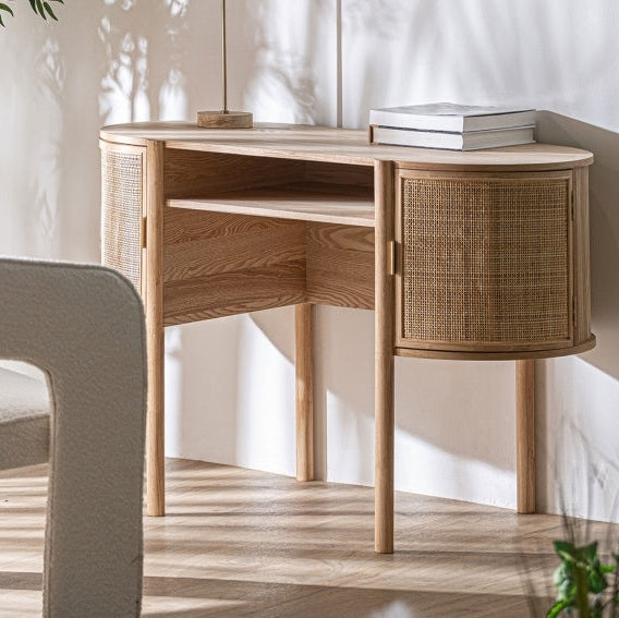 El escritorio nórdico en madera de mango y ratán Ikla, de diseño minimalista y puertas de ratán curvas, se apoya en una pared blanca. Dos libros y una lámpara reposan sobre él, mientras la luz del sol proyecta sombras en el suelo y la pared.